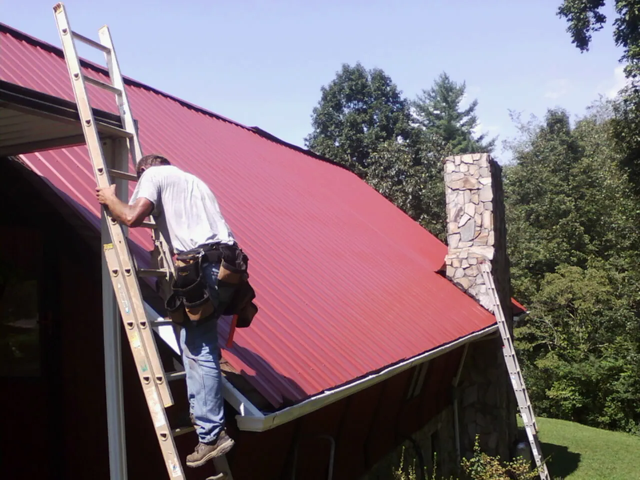 Roofer on a ladder beside a red metal roof and stone chimney — Soffit & Fascia Repair by Roofing Brothers in Scranton